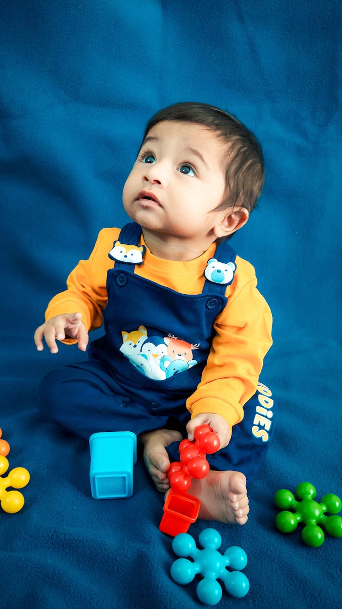 About Cute baby sitting barefoot in colorful overalls, surrounded by toys in a studio setting.