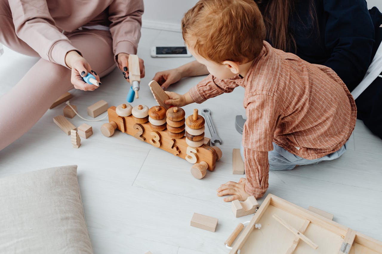 About A young child engaged with a wooden toy train in a warm indoor setting, surrounded by adults.
